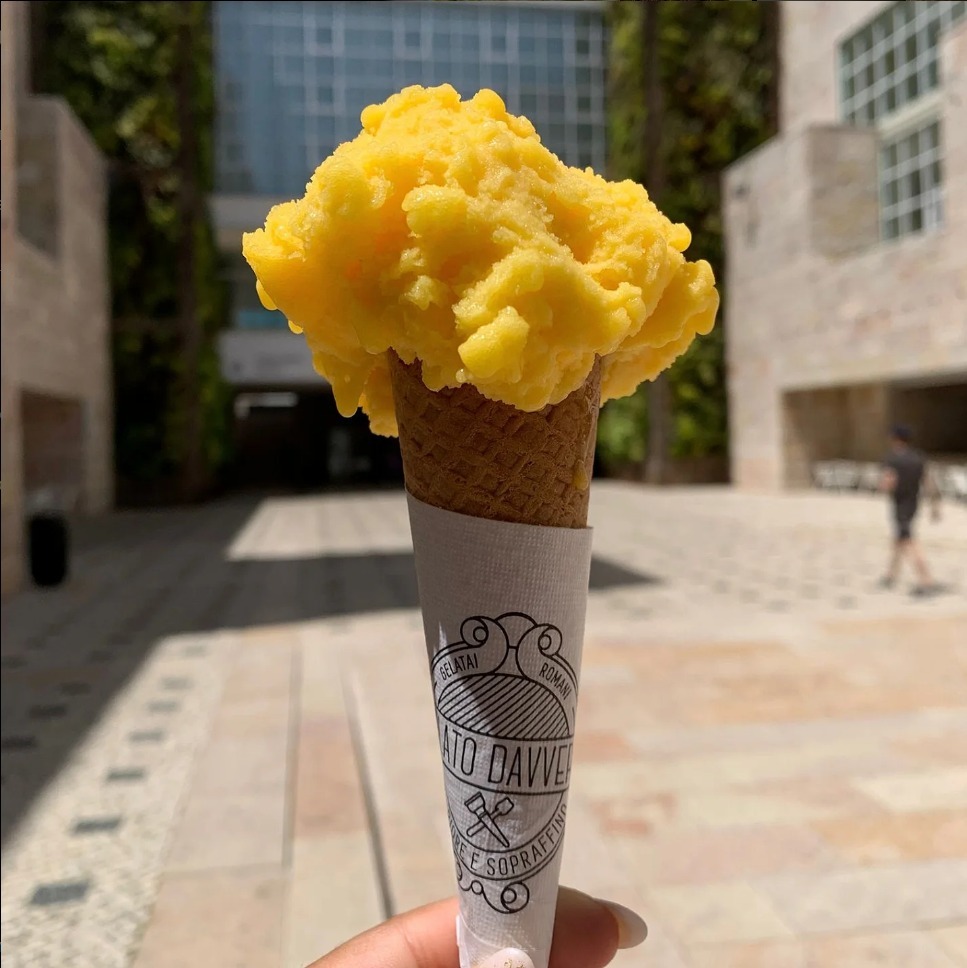 Close-up of a vibrant yellow gelato in a waffle cone held outdoors, with modern architecture in the background.