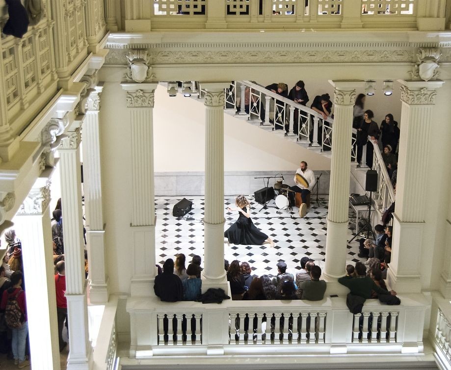 Dance performance in grand hall with pillars, musician playing drums, and an audience watching from balcony.
