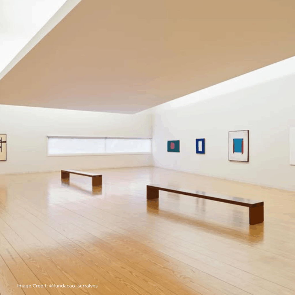 Modern art gallery interior featuring wooden benches and abstract paintings on white walls. Bright natural light streams in.