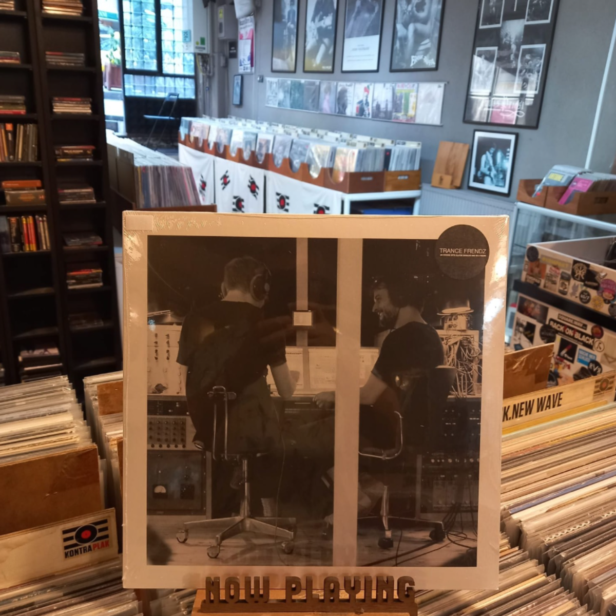 Vinyl records on display in a music store with 'Now Playing' sign and two DJs engaged in a live session.