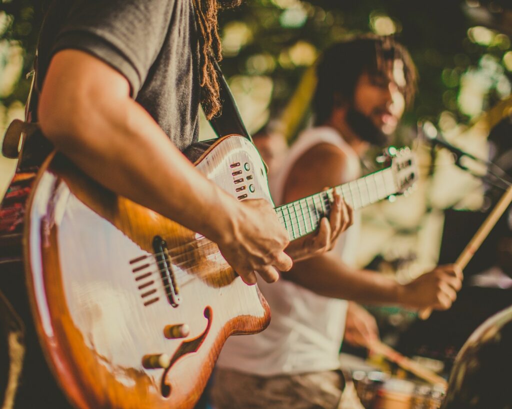 Musicians performing live, showcasing a guitarist playing a wooden guitar with a drummer in the background.