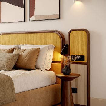 Modern bedroom with mustard headboard, abstract art, and wooden side table with a vase, lit by a wall sconce.