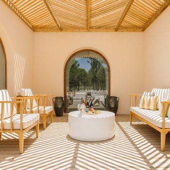 Ando Living Comporta Villas. Cozy outdoor lounge with wooden furniture, bright cushions, and a round table, bathed in sunlight and shadow patterns.