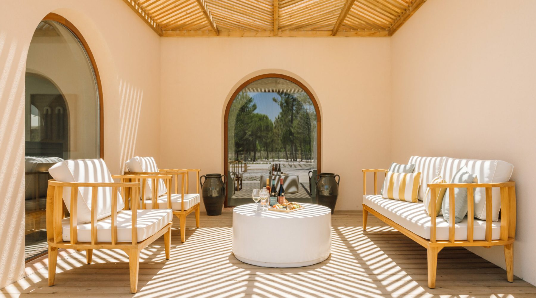 Ando Living Comporta Villas. Cozy outdoor lounge with wooden furniture, bright cushions, and a round table, bathed in sunlight and shadow patterns.