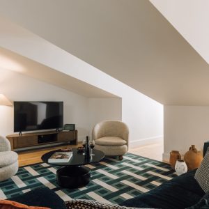 Cozy attic living room with TV, textured armchairs, geometric rug, and warm lighting under a sloped ceiling.