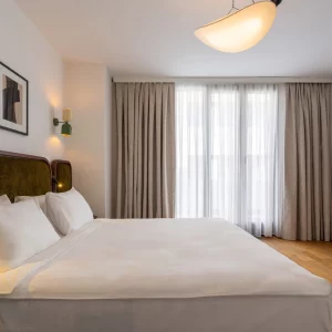 Modern bedroom featuring a stylish bed, elegant decor, and large windows with sheer curtains. Cozy and inviting ambiance.